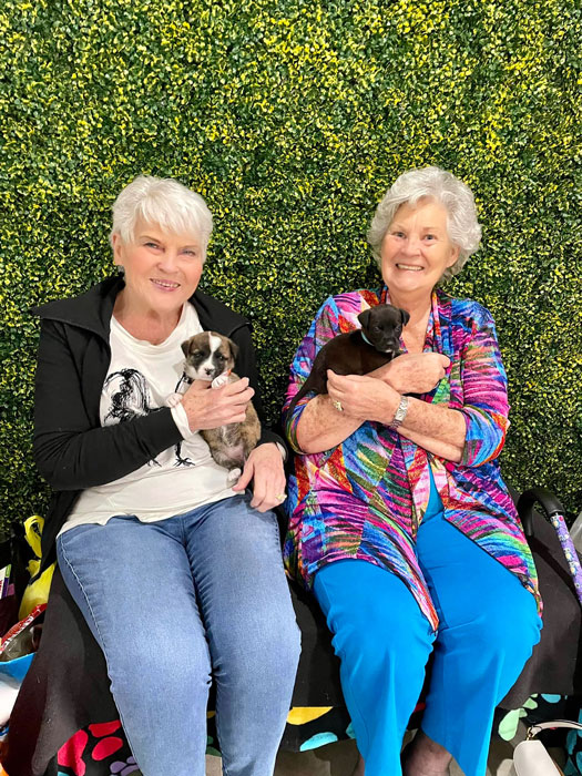 Two senior residents sitting together, each holding a small puppy, against a green leafy background, showcasing the joy and companionship of pet therapy in senior living.