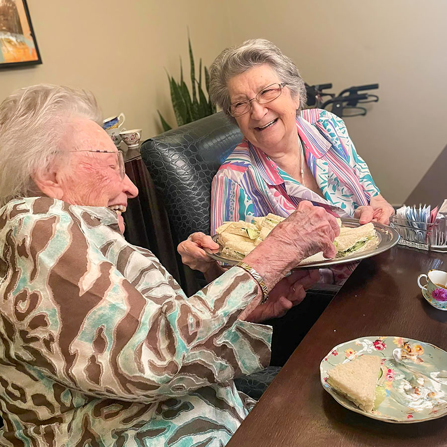 Two senior residents share a joyful moment at a tea party, laughing together as they hold a platter of finger sandwiches, with floral plates and teacups on the table in front of them.