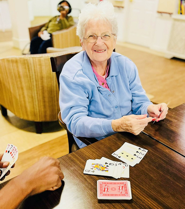 Senior resident enjoying a game of cards, smiling warmly while sitting at a table with playing cards spread out, engaged in a lively game in a cozy senior living community.
