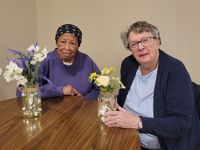 Two senior residents sitting at a table, smiling. One wears a black headscarf and purple shirt, while the other wears glasses and a blue cardigan. Vases of flowers are on the table.