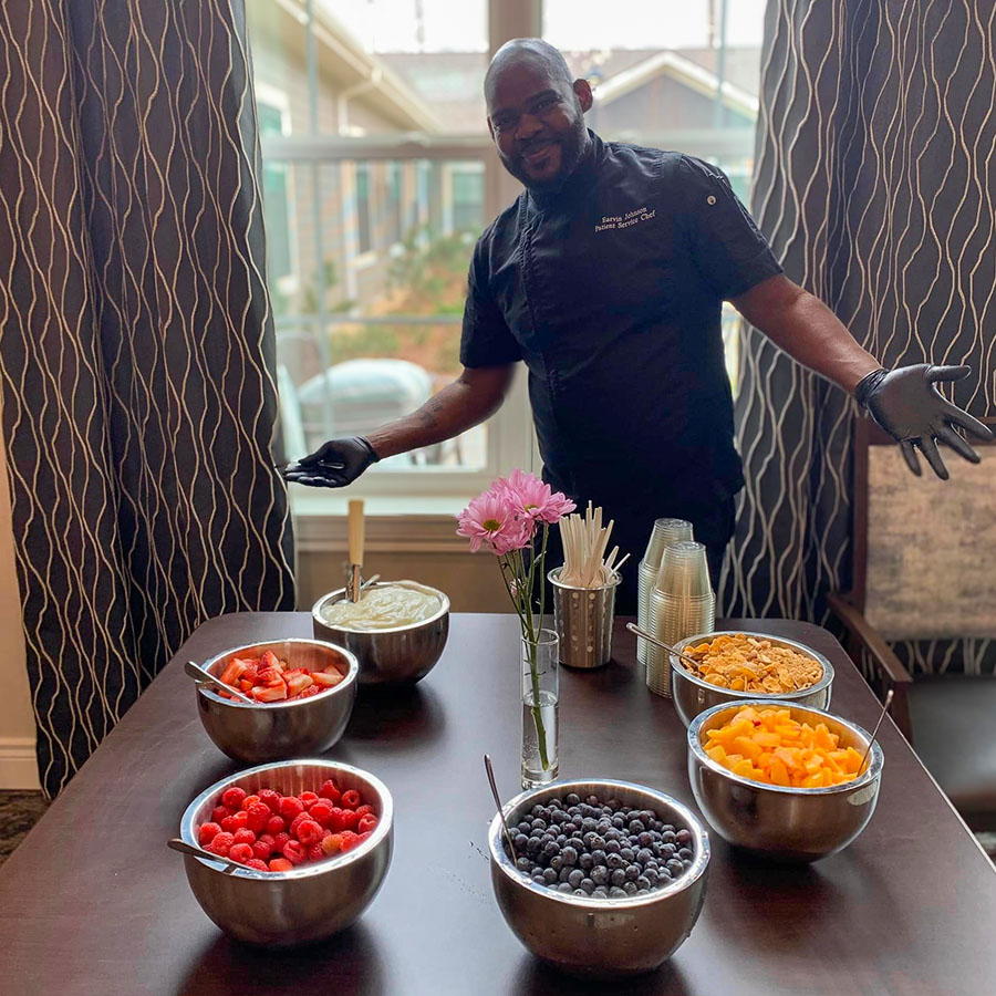 Chef presenting a colorful yogurt bar setup with bowls of fresh fruits, including strawberries, raspberries, blueberries, and peaches, along with granola and yogurt options.