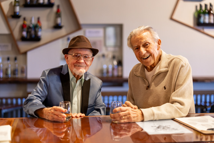 Two senior residents enjoy drinks together at a bar, smiling and holding glasses, dressed in casual and formal attire during a social gathering at Vitalia Senior Living.