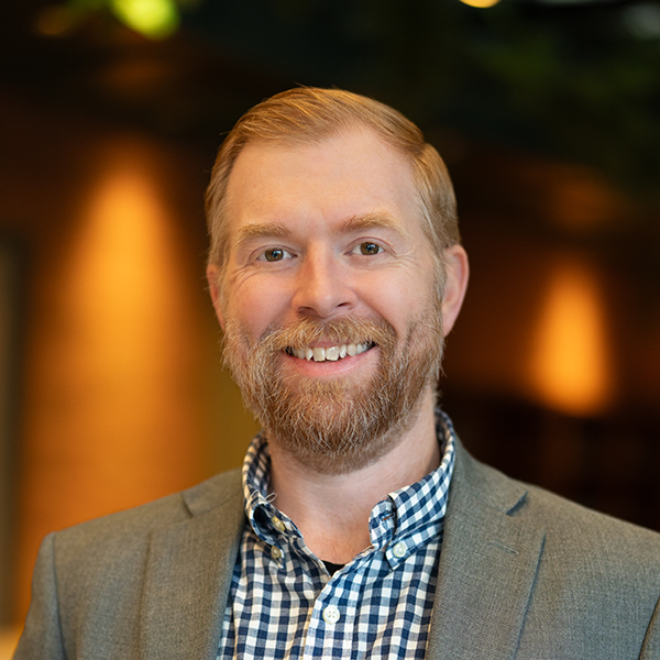 Adam Zanger, Executive Director at Curtis Creek Senior Living, smiling confidently while wearing a gray blazer and checkered shirt, standing in a warmly lit setting.
