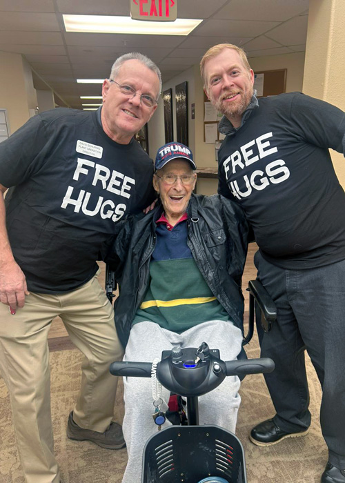 Two team members wearing 'Free Hugs' shirts pose with a smiling senior resident sitting in a mobility scooter inside the senior living community.