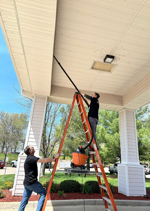 Two team members clean hard-to-reach exterior surfaces with a vacuum on a tall ladder under a covered entryway during spring cleaning.