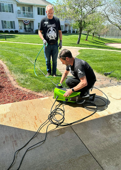 Two team members prepare a pressure washer to clean an outdoor walkway surrounded by fresh-cut grass and landscaping at a senior living community.