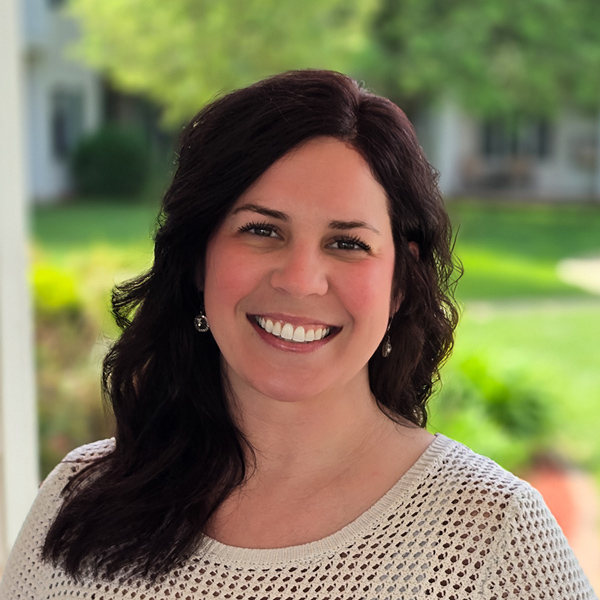 Christy Wellman, Senior Living Director at Curtis Creek Senior Living, smiling warmly outdoors in a cream knit top with a lush, green background behind her.