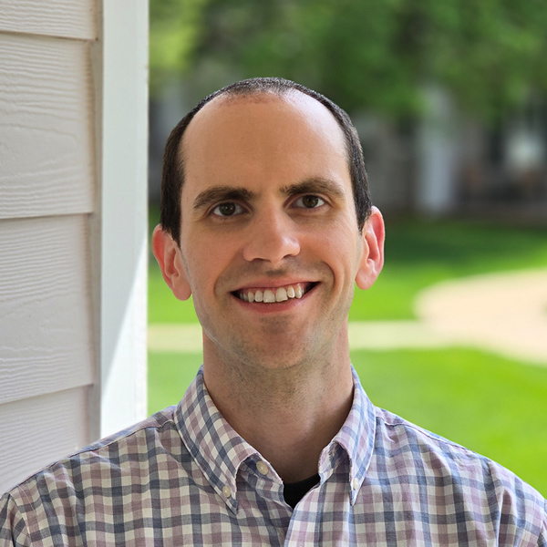 Mitchell Provow, Senior Living Director at Curtis Creek Senior Living, smiling outdoors in a checkered shirt with a sunny green lawn and walking path in the background.