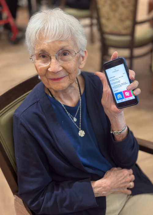 A smiling senior woman with short white hair and glasses holds up a smartphone displaying the Arrow app while seated in a dining area.