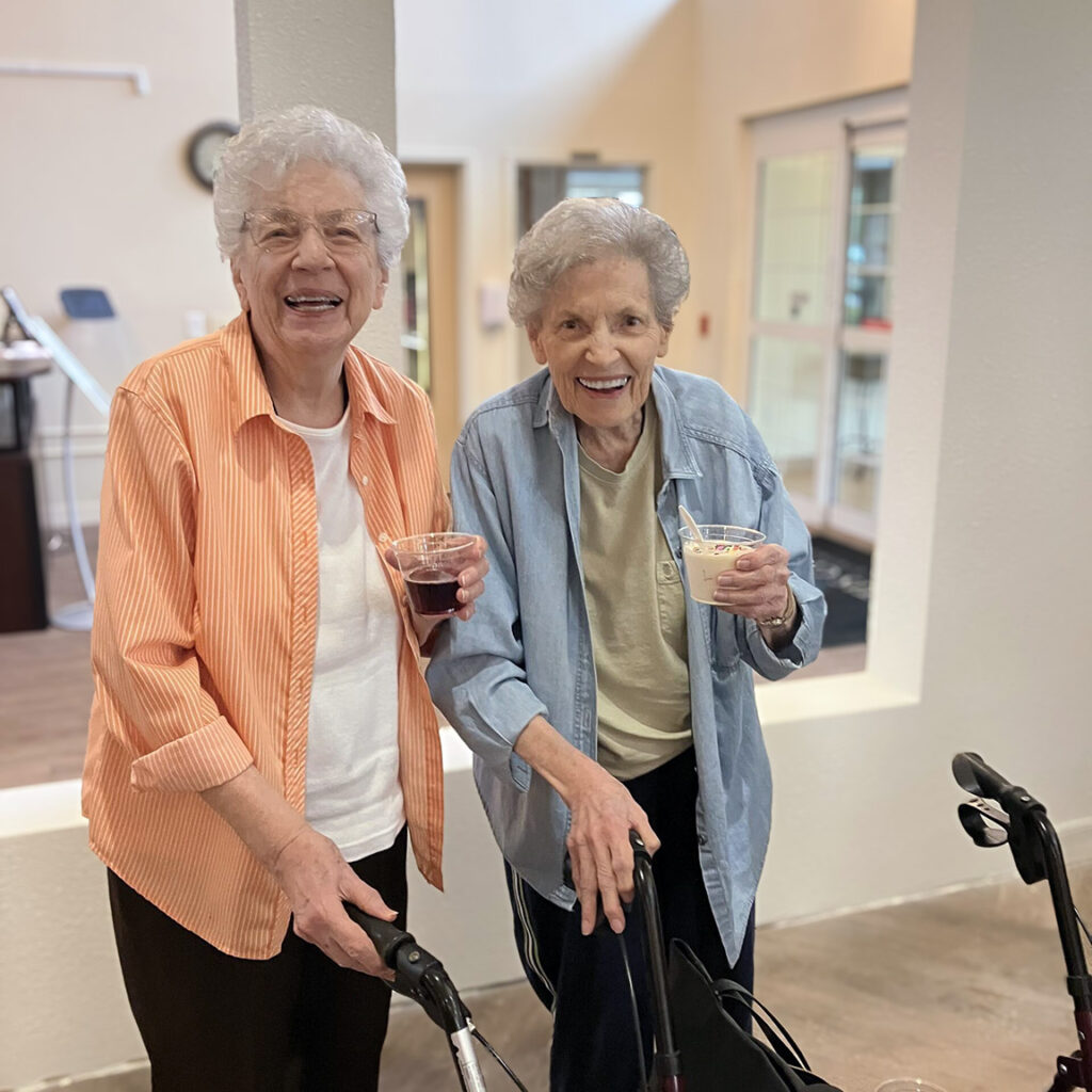 Two smiling senior women stand side by side, happily holding beverages.
