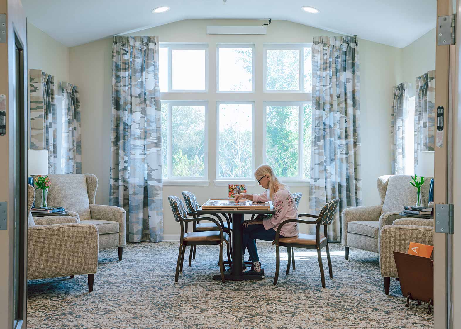 Wide shot of a senior woman with long, blonde hair seated in a room with high ceilings, windows all the way up on the far wall, and lots of bright, natural light shining off of soft white and beige toned surfaces.