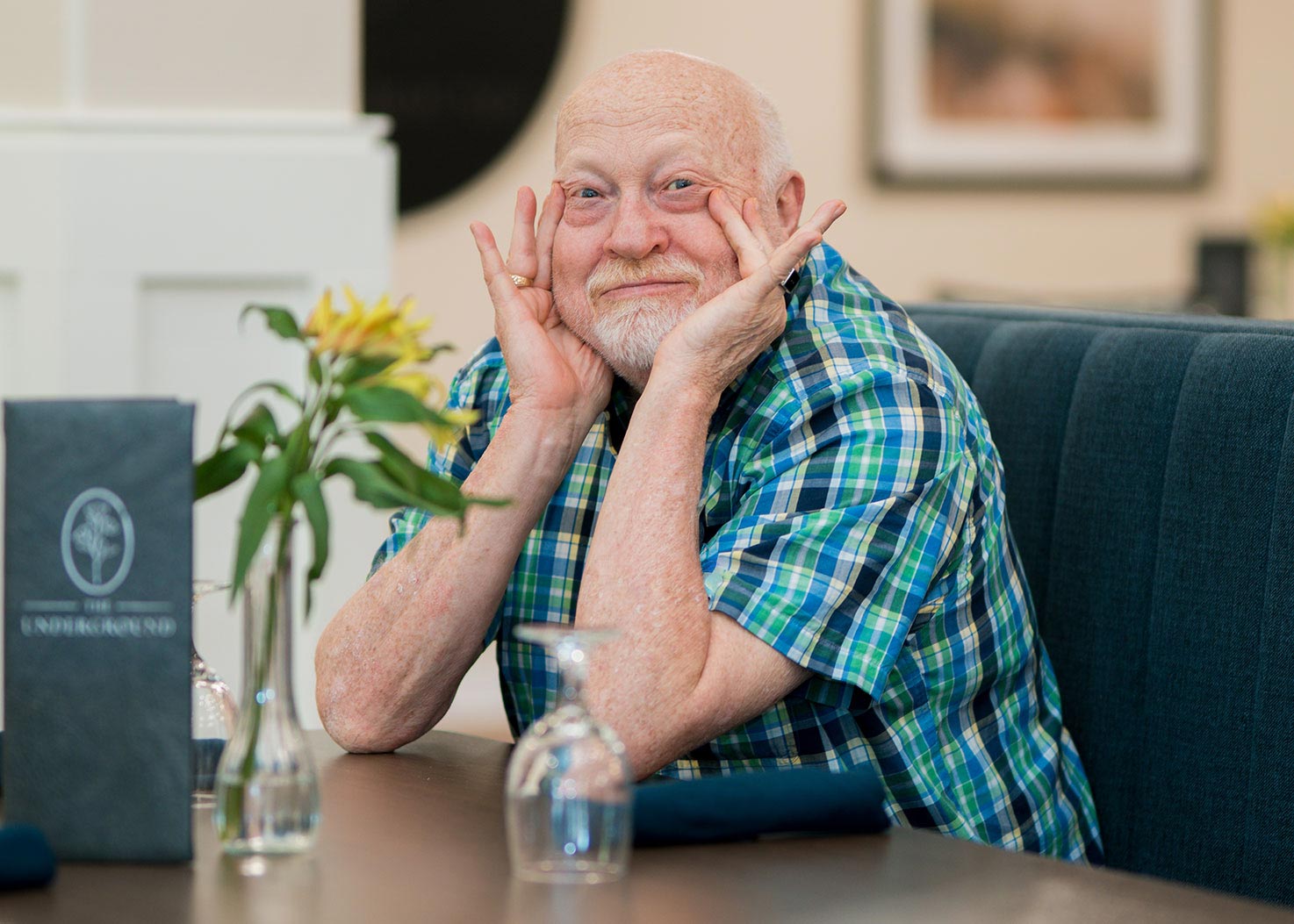 A smiling senior man holds his hands on his cheeks playfully while sitting at a dining table with a menu for Curtis Creek's on-site restaurant, The Underground.