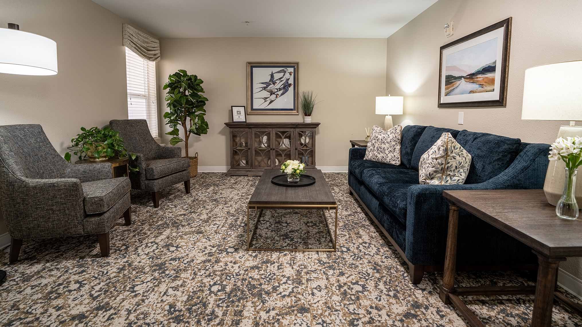 A cozy senior living lounge with a navy sofa, patterned rug, two gray chairs, and warm lighting, creating a comfortable and inviting space for relaxation and conversation.