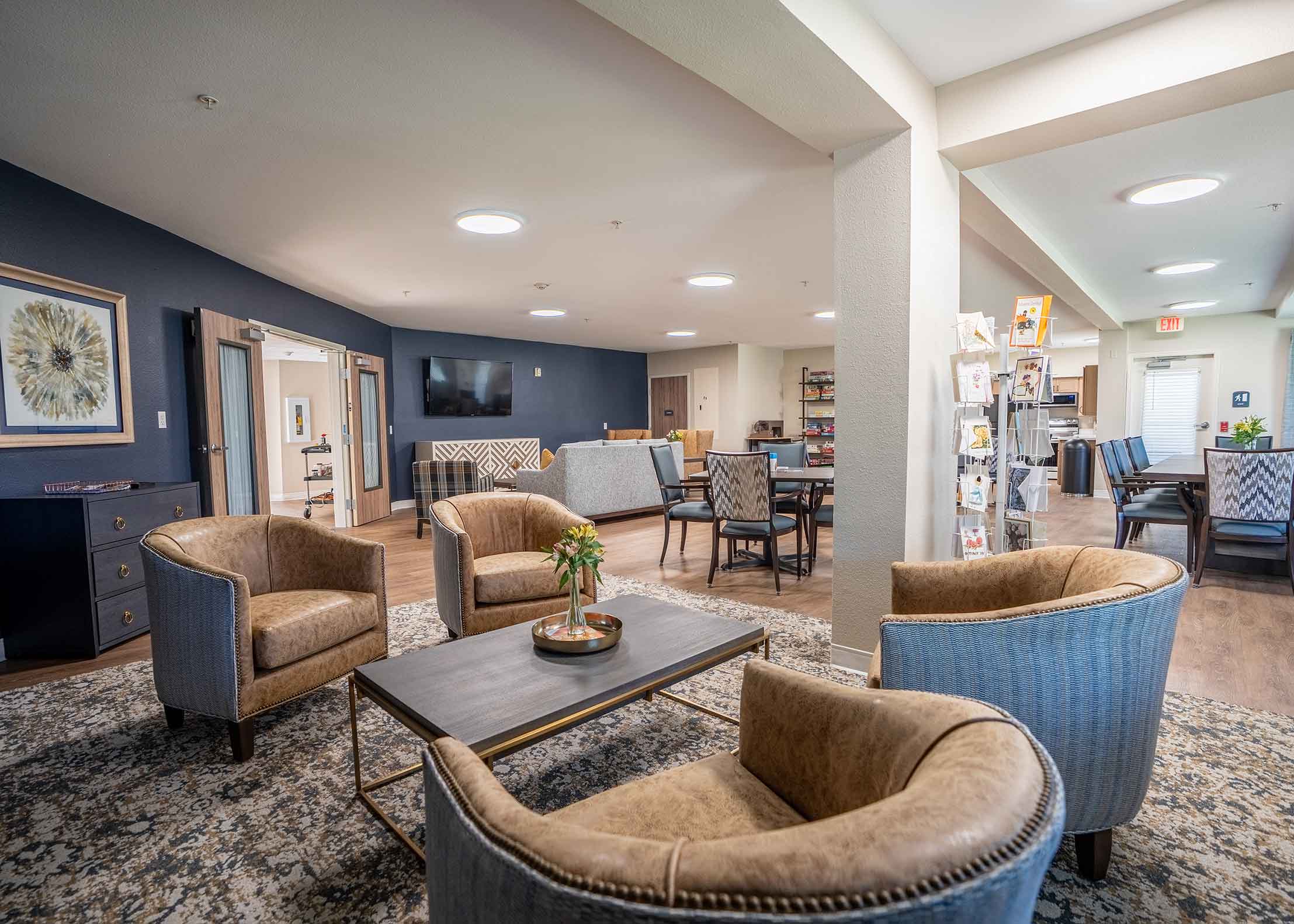 A spacious senior living lounge featuring comfortable seating, a coffee table with flowers, dining tables, and a television, creating a warm and inviting community space.