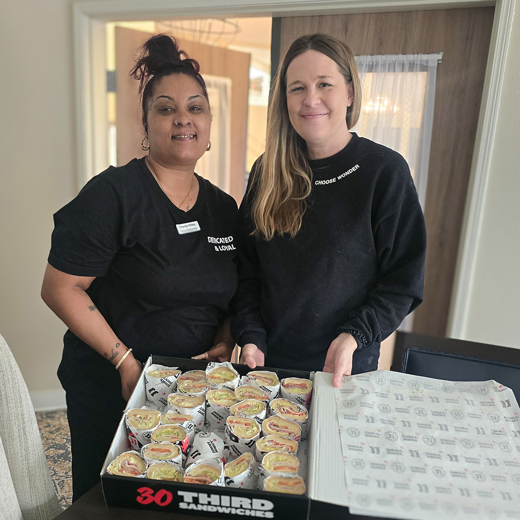 Two Curtis Creek Senior Living team members smile with a large box of sandwiches.