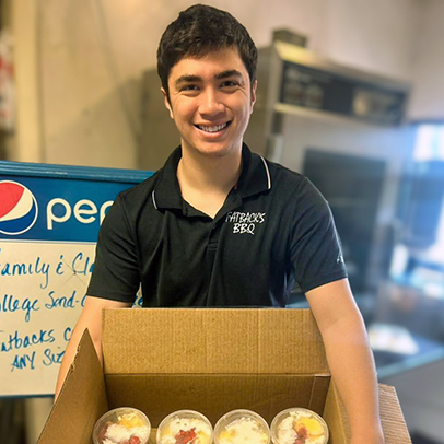A young man smiles, holding a box of treats, at a local barbecue establishment.