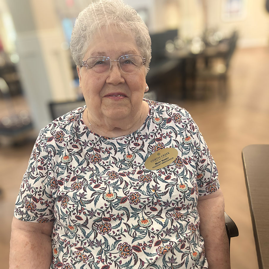 A senior woman wearing a Resident Ambassador badge smiles at Curtis Creek Senior Living, ready to help new residents feel at home.